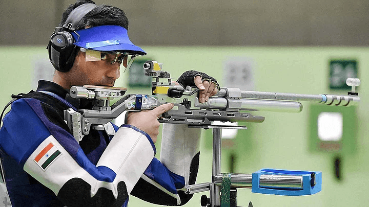 A shooter in a blue jacket and cap aiming a match air rifle