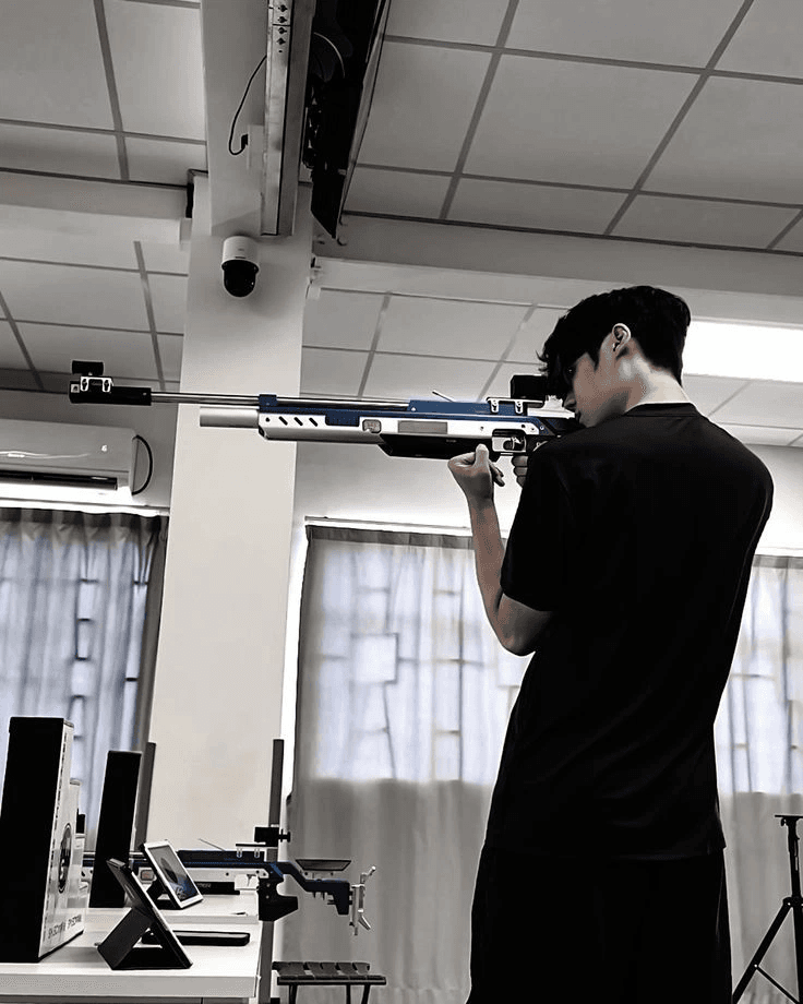 A shooter standing on the line with a match air rifle raised, seen from behind and to the side